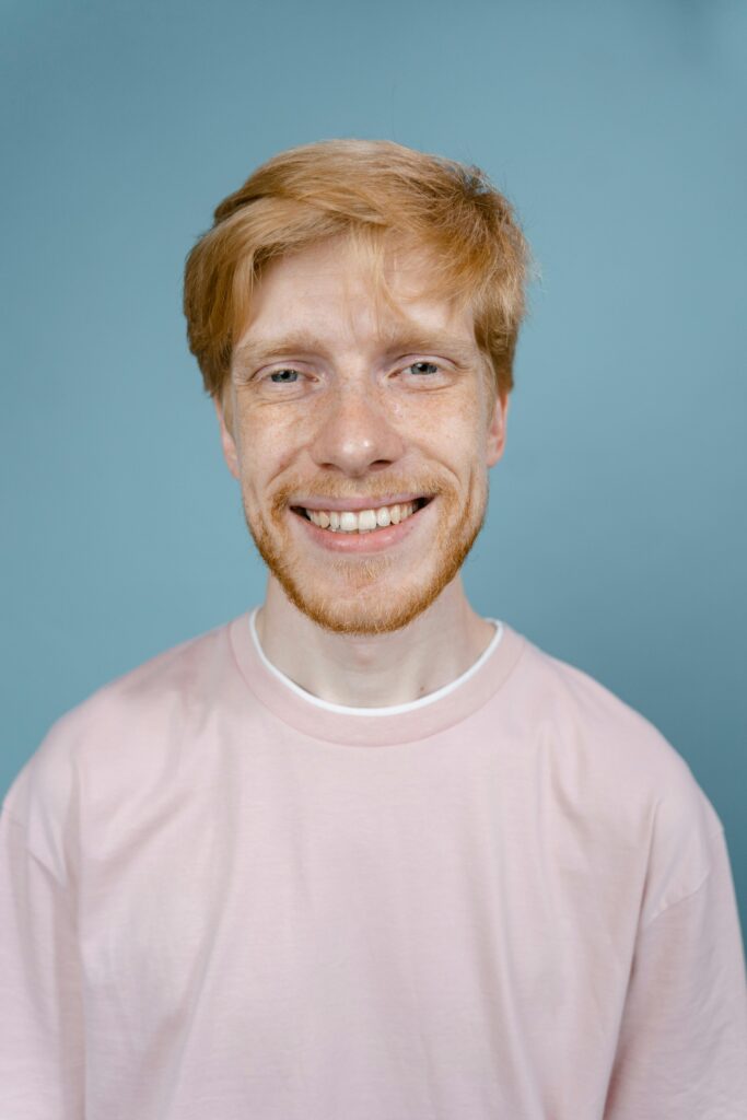 Portrait of a smiling bearded man with blonde hair wearing a pink shirt in studio setting.
